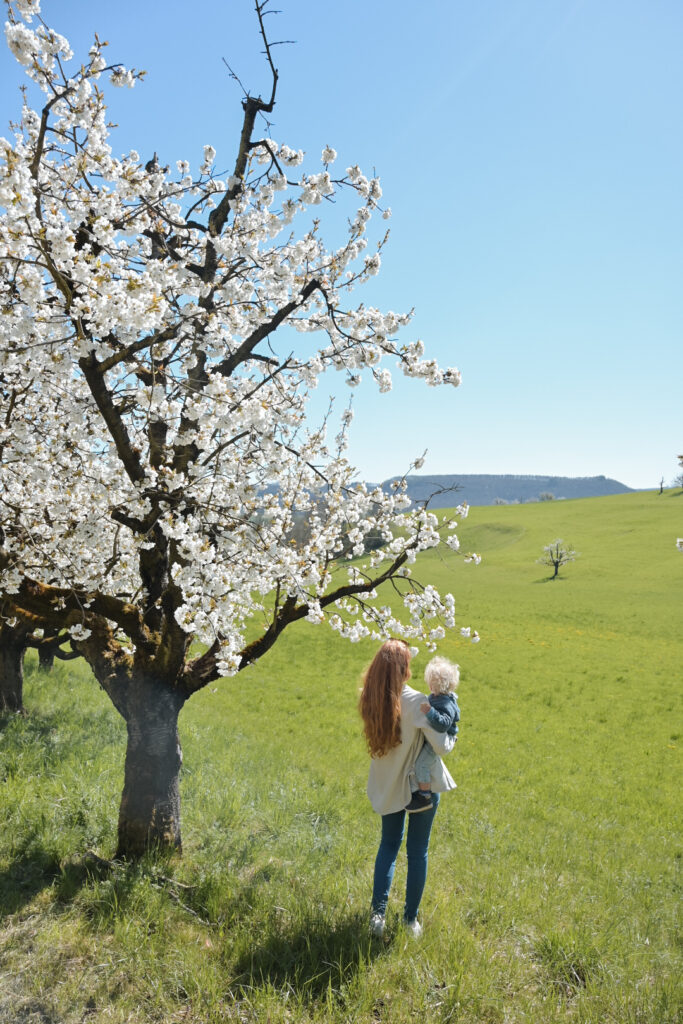 Foto von Frau mit Baby auf dem Arm unter Kirschbaum schaut ins Tal